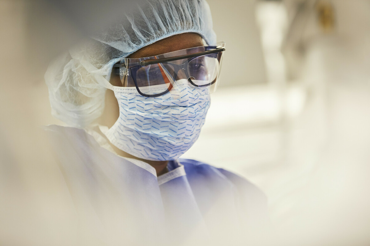 Person in scrubs with hairnet, eye shield over glasses, and a surgical mask